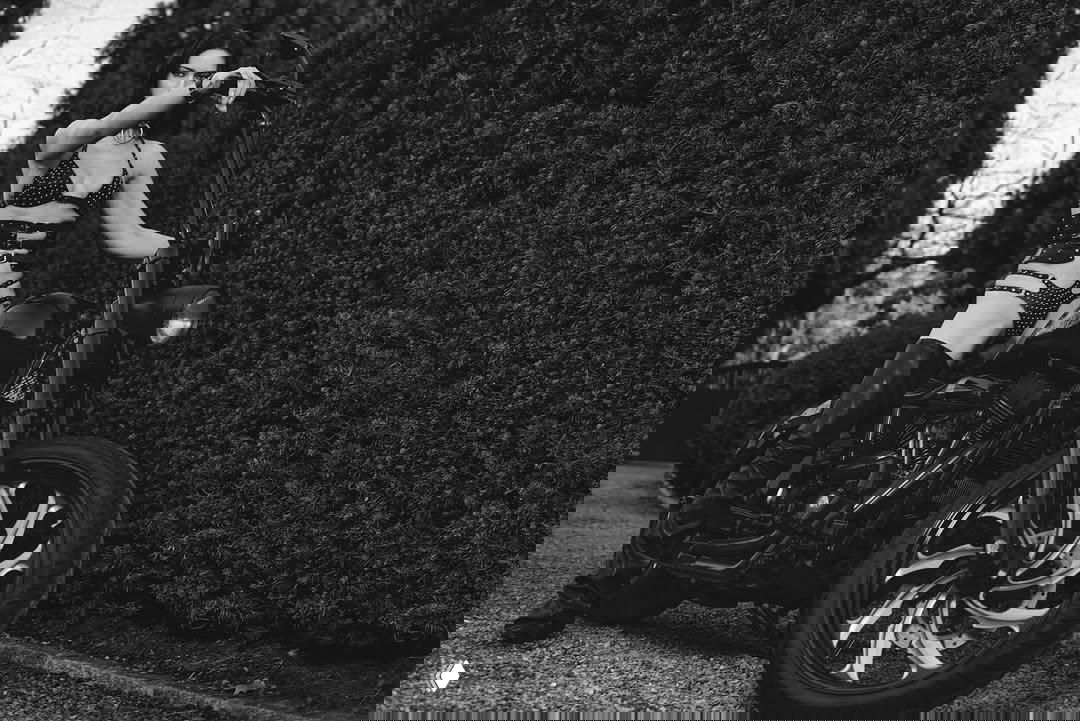 Черно‑белый крупный кадр: женщина в откровенном комплекте сидит на мотоцикле у живой изгороди, акцент на силуэте и деталях мотоцикла.