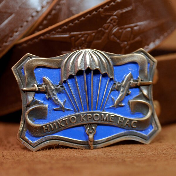 Металлический шильд с парашютной эмблемой и надписью «НИКТО КРОМЕ НАС» на фоне ремней — отдельный крупный значок/медальон рядом с изделиями.