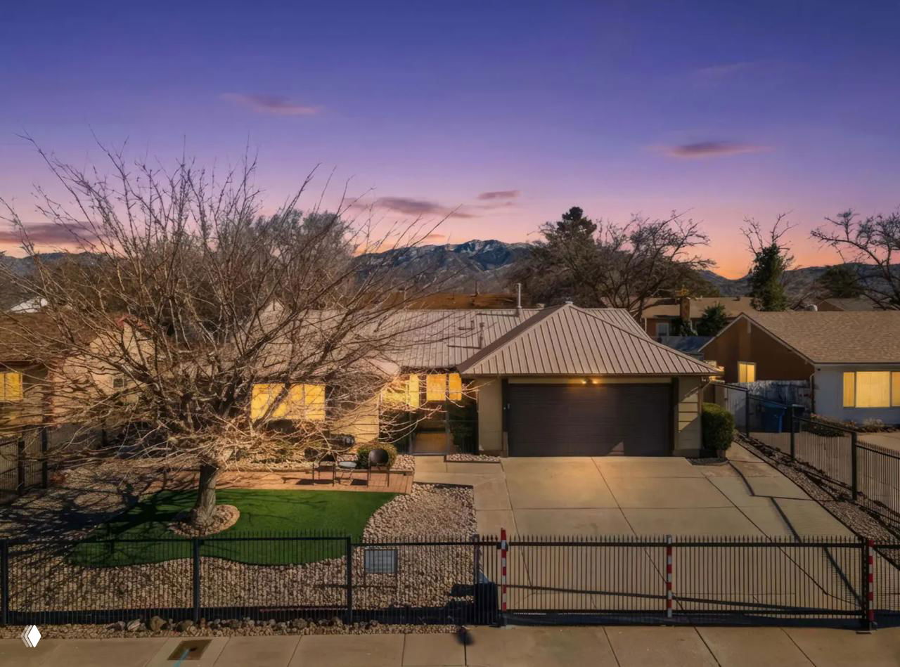 Вечерний фасад дома в Альбукерке: одноэтажная резиденция с гаражом, огороженным двором, освещёнными окнами и горами на заднем плане.