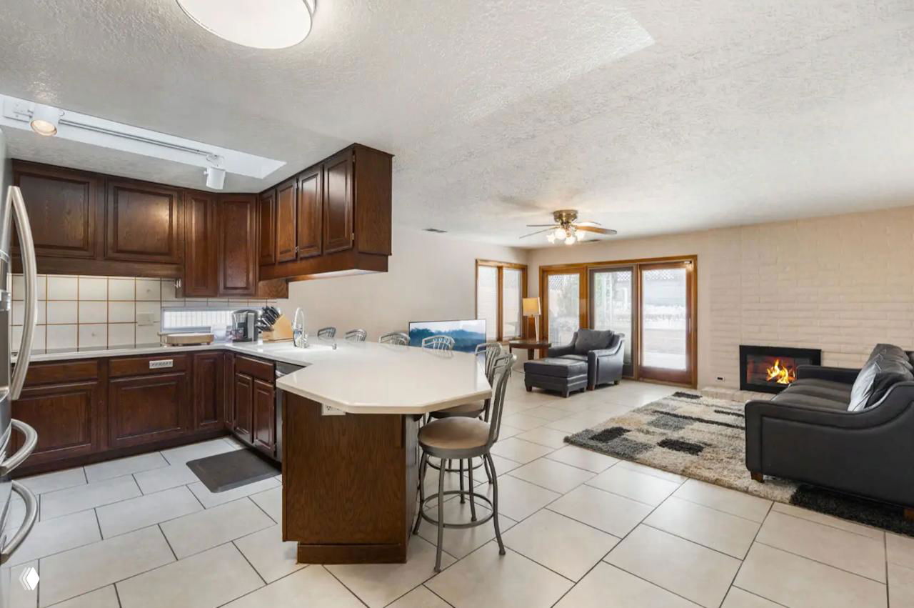 Интерьер дома: открытая кухня со стойкой и деревянными шкафами, плиточный пол и гостиная с камином, вид на зону отдыха и столовую часть.