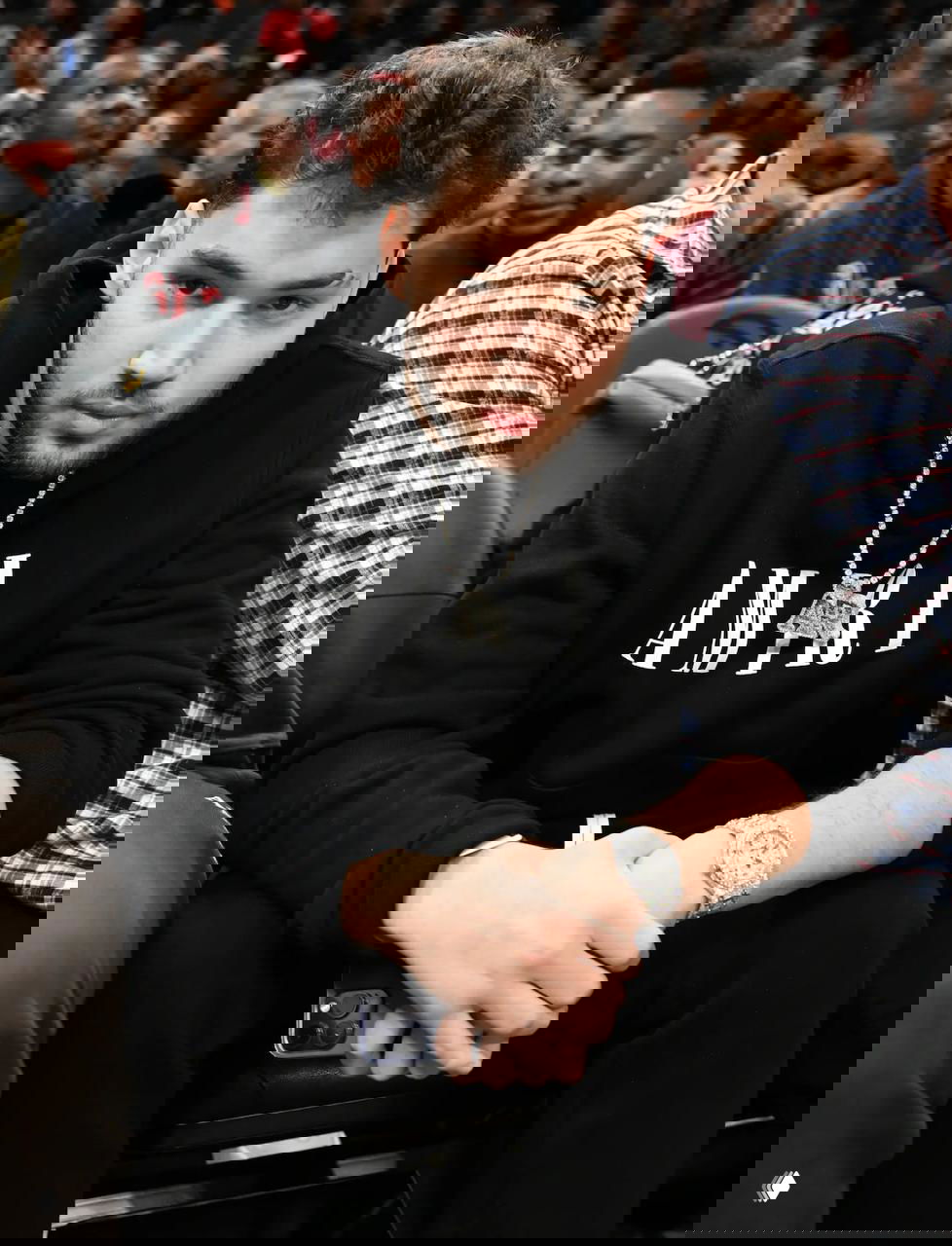 Молодой мужчина в чёрном худи и с крупной серебряной цепочкой сидит на трибуне во время баскетбольного матча, вокруг зрители на трибунах.