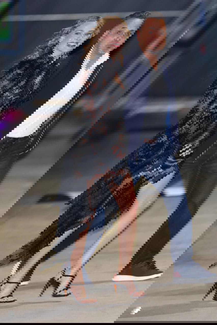 Фотоснимок женщины в длинном прозрачном кружевном платье, улыбается и идёт рядом с сопровождающим на фоне городского пейзажа