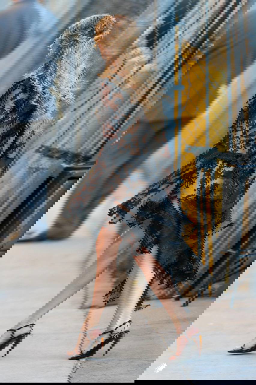 Женщина в чёрном ажурном платье идёт по тротуару, запечатлён момент сзади, видны волнистые волосы и движение ткани