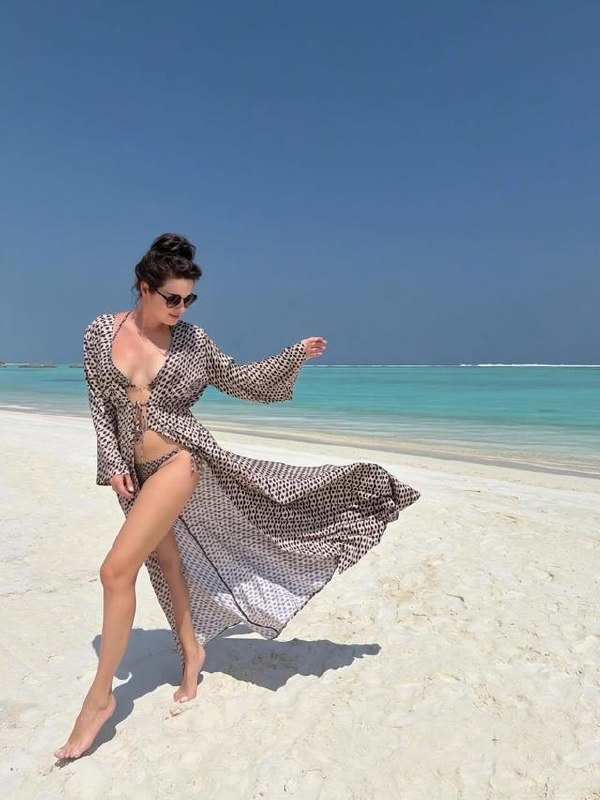 Модель в пляжной тунике и бикини шагает по белому песку у бирюзового моря с ясным горизонтом, летняя курортная фотография.