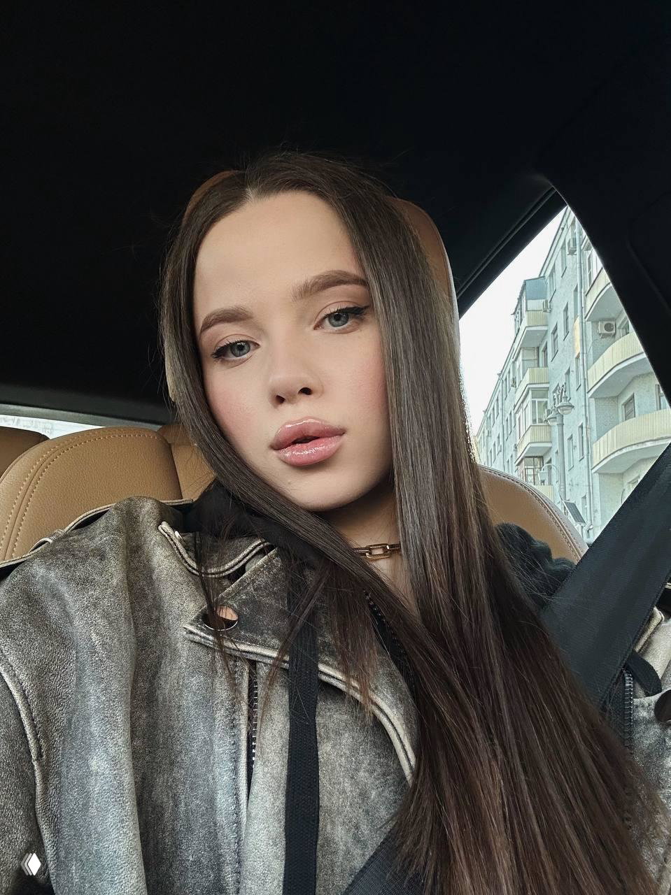 Селфи молодой женщины в автомобиле: лицо крупным планом, макияж, прямые тёмные волосы, на заднем плане городская улица и фасады зданий.