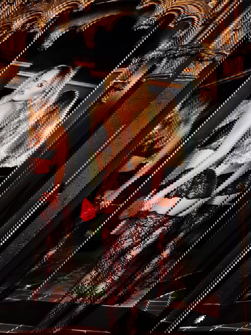 Женщина в кружевном чёрном нижнем белье и чулках позирует перед зеркалом в роскошной студии — кадр из рекламной съёмки.