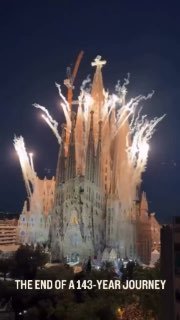 Вертикальное ночное фото верхней части Саграда Фамилия: освещённый крест, новая завершённая башня и фейерверки.