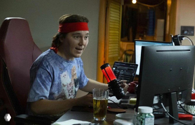 Кадр из фильма: харизматичный видеоблогер за столом с микрофоном и мониторами, на столе стакан с напитком и предметы быта, сцена стрим‑обстановки.