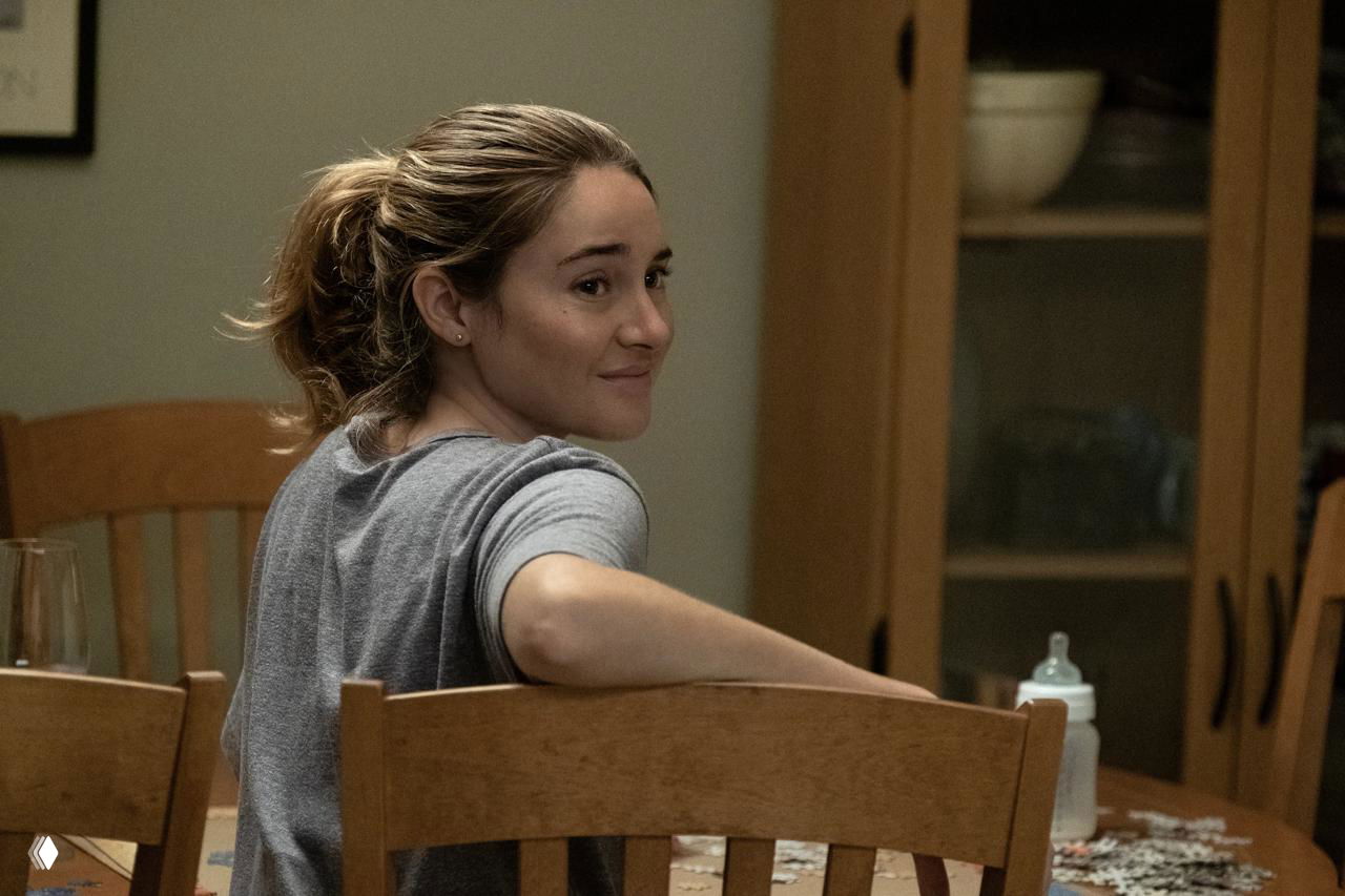 Бытовой кадр с женщиной за кухонным столом: героиня улыбается, на столе детская бутылочка и пазл, подчёркивающие семейную сторону персонажей в сюжете.