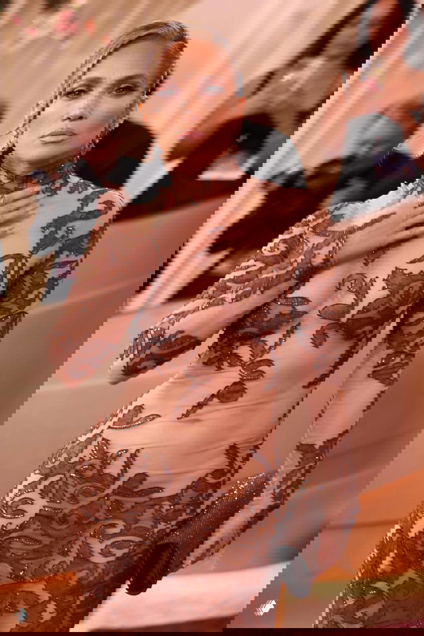 Крупный план женщины в облегающем бежево-коричневом платье с узорной отделкой на фоне красной дорожки и декора Golden Globes.