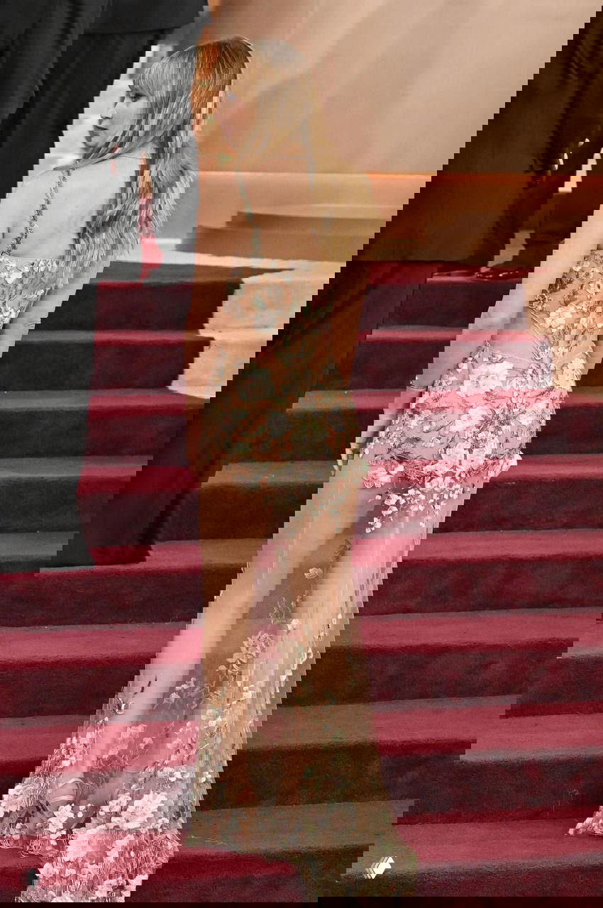 Кадр со спины: женщина в длинном полупрозрачном платье с цветочной вышивкой поднимается по ступеням красной дорожки на церемонии 2026 года.