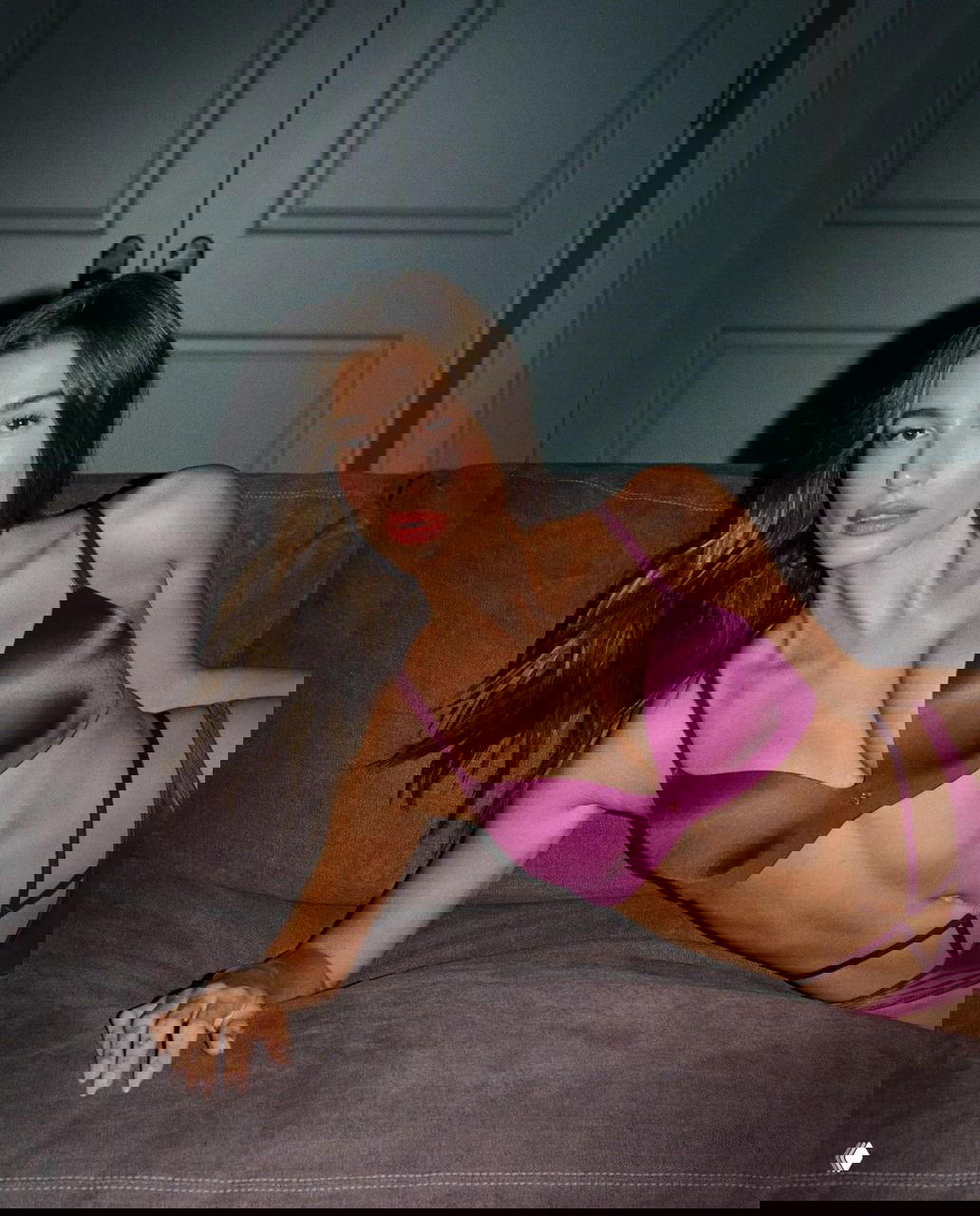 Крупный портрет женщины в пурпурном бюстье на диване, вечернее освещение, слегка лежащая поза, фокус на верхней части тела, серия фотографий высокого разрешения.