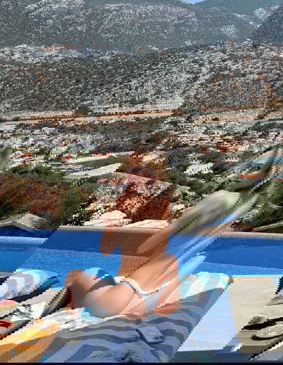 Модель сидит на шезлонге у края бассейна в белом купальнике, демонстрируя осанку и спину на фоне гор и домов курорта.