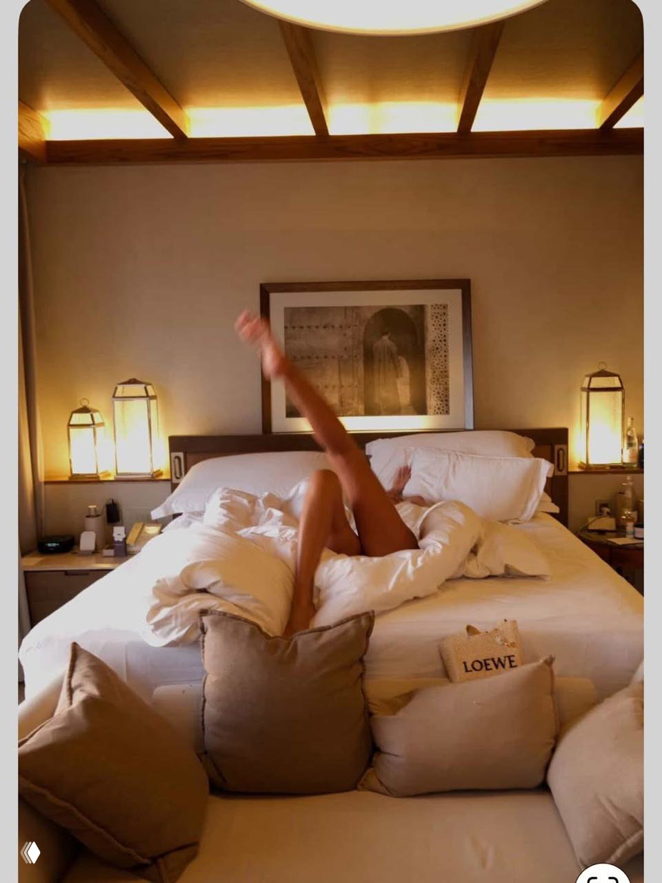 Интерьерный кадр: женщина в постели под одеялом с поднятой ногой в комнате отеля; тёплое освещение и аккуратная декоративная обстановка на заднем плане.