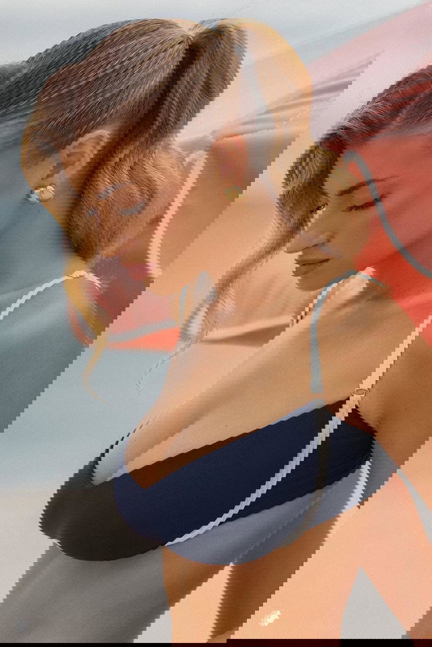 Крупный план Фрейи Тайди в темно‑синем купальнике у пляжного зонта, натуральный макияж и поза, подчёркивающая линию плеч и декольте.