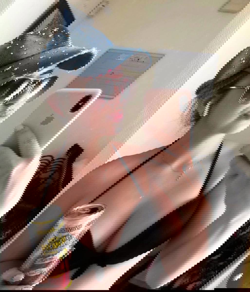 Молодая женщина в чёрном купальнике и бейсболке делает селфи в зеркале, держа в руке банку напитка — видна часть номера и аксессуары.