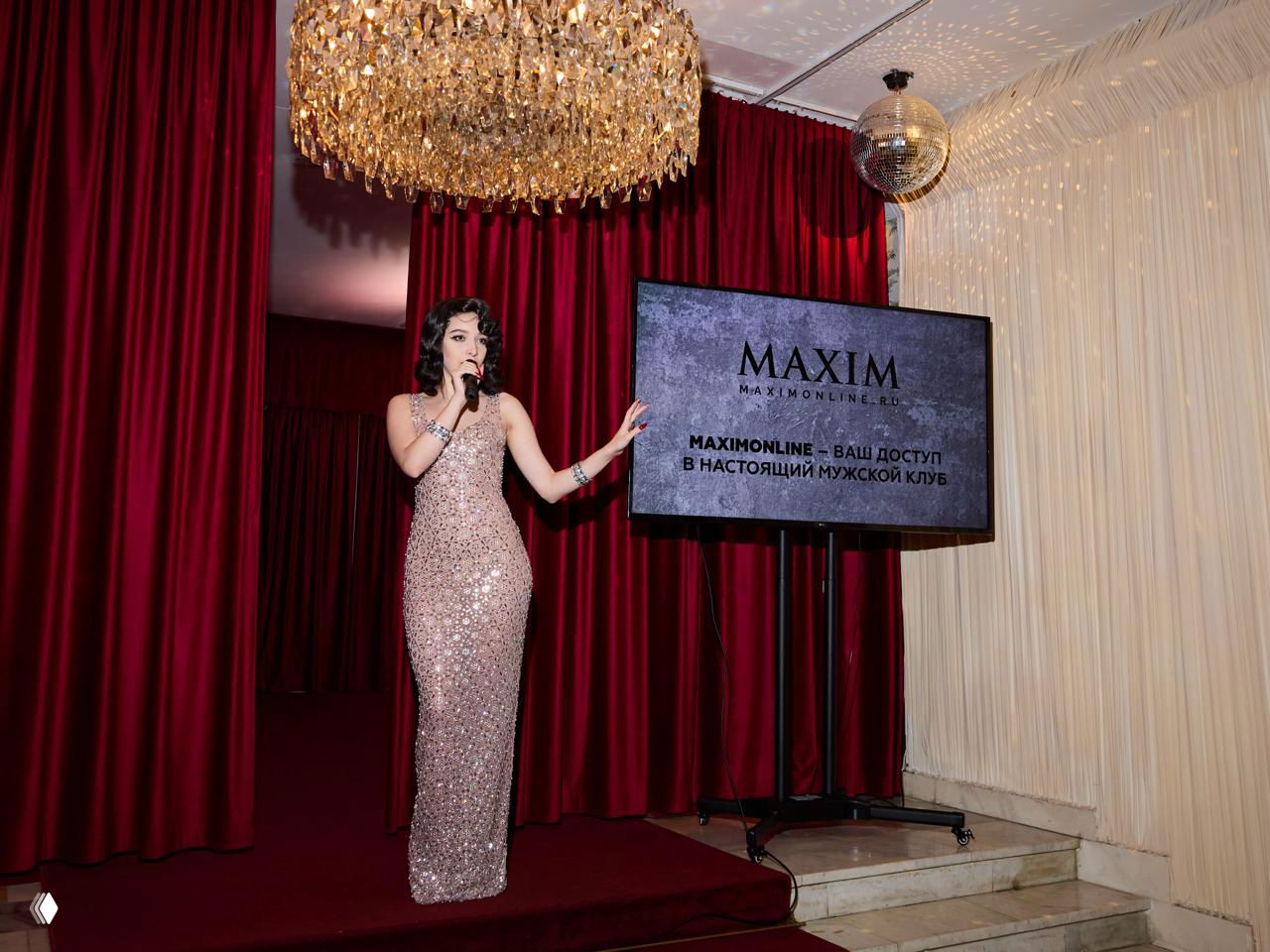 Артистка в длинном блестящем платье на сцене рядом с экраном Maxim, кадр показывает интерьер зала с люстрой и сценой презентации
