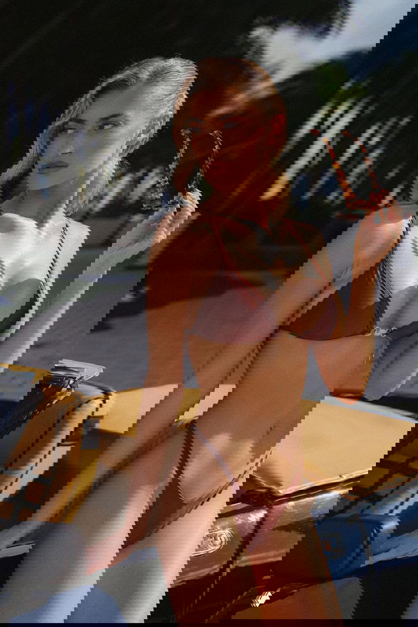 Фрейя Тайди в коричневом бикини стоит рядом с кабриолетом, солнце подчёркивает контуры фигуры на фоне зелёных пальм и тропического сада.