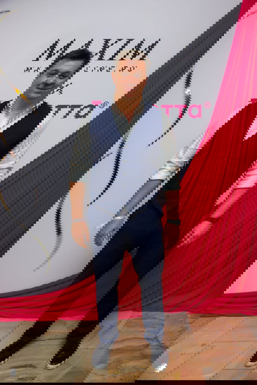 Мужчина в жилете и брюках стоит на фоне брендированной стены Maxim, кадр с фотозоны показывает плитку пола и декоративную драпировку