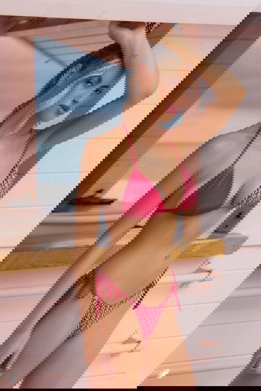 Фрейя Тайди в розово‑красном бикини у пляжной будки на побережье, лёгкая поза и тёплое освещение с видом на море.