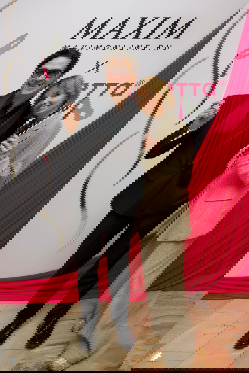 Двое гостей в элегантных костюмах позируют на фоне баннера Maxim и яркой розовой драпировки, фото в стиле красной ковровой фотозоны