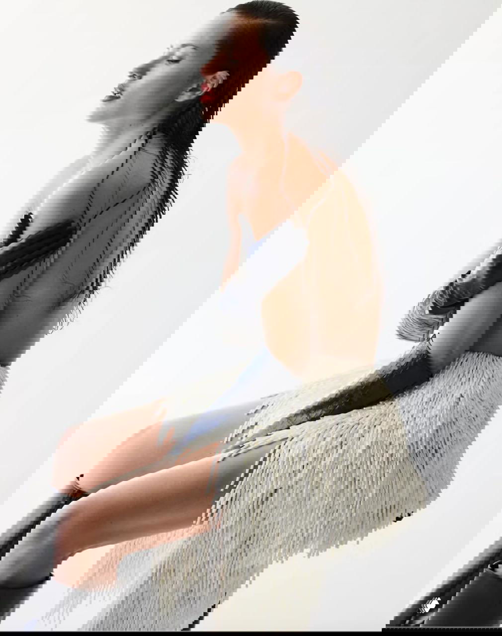 Профильный снимок Адрианы Лимы из фотосессии Harper’s Bazaar: мокрые волосы, длинные перчатки и текстурированная юбка с бахромой, чувственная поза на светлом фоне.