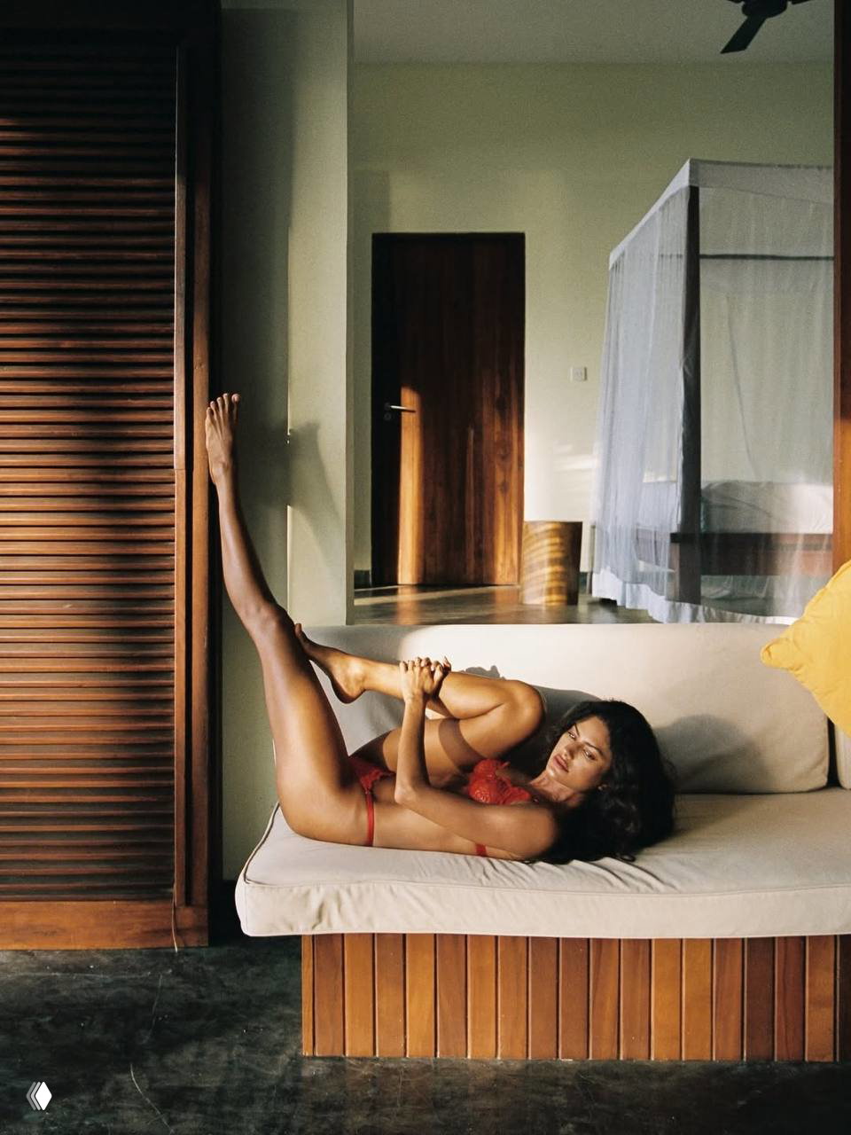 Бразильская модель в красном кружевном белье лежит на диване в уютном интерьере тропического отеля при тёплом утреннем свете.