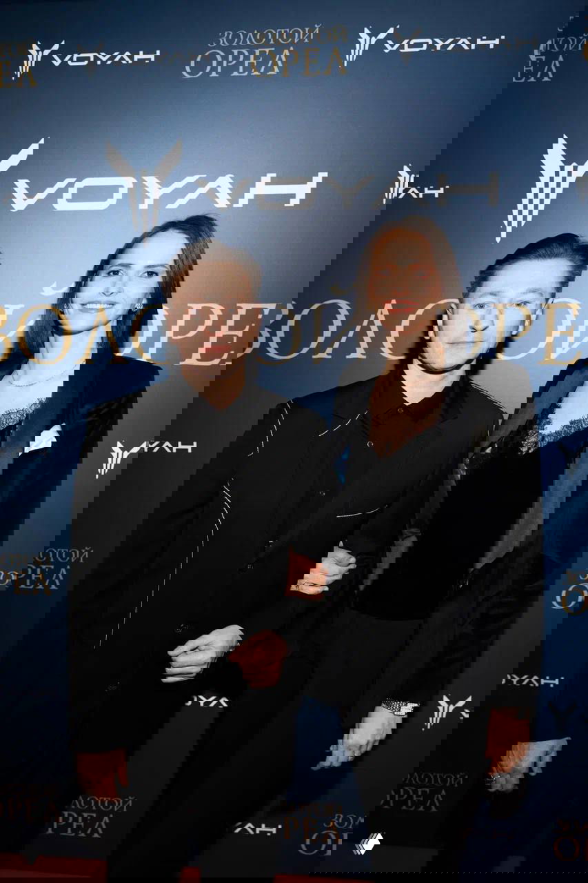 Гости на пресс-воле церемонии «Золотой орёл» позируют на фоне бренд-стены партнёра VOYAH; двое взрослых в вечерних нарядах смотрят в камеру.