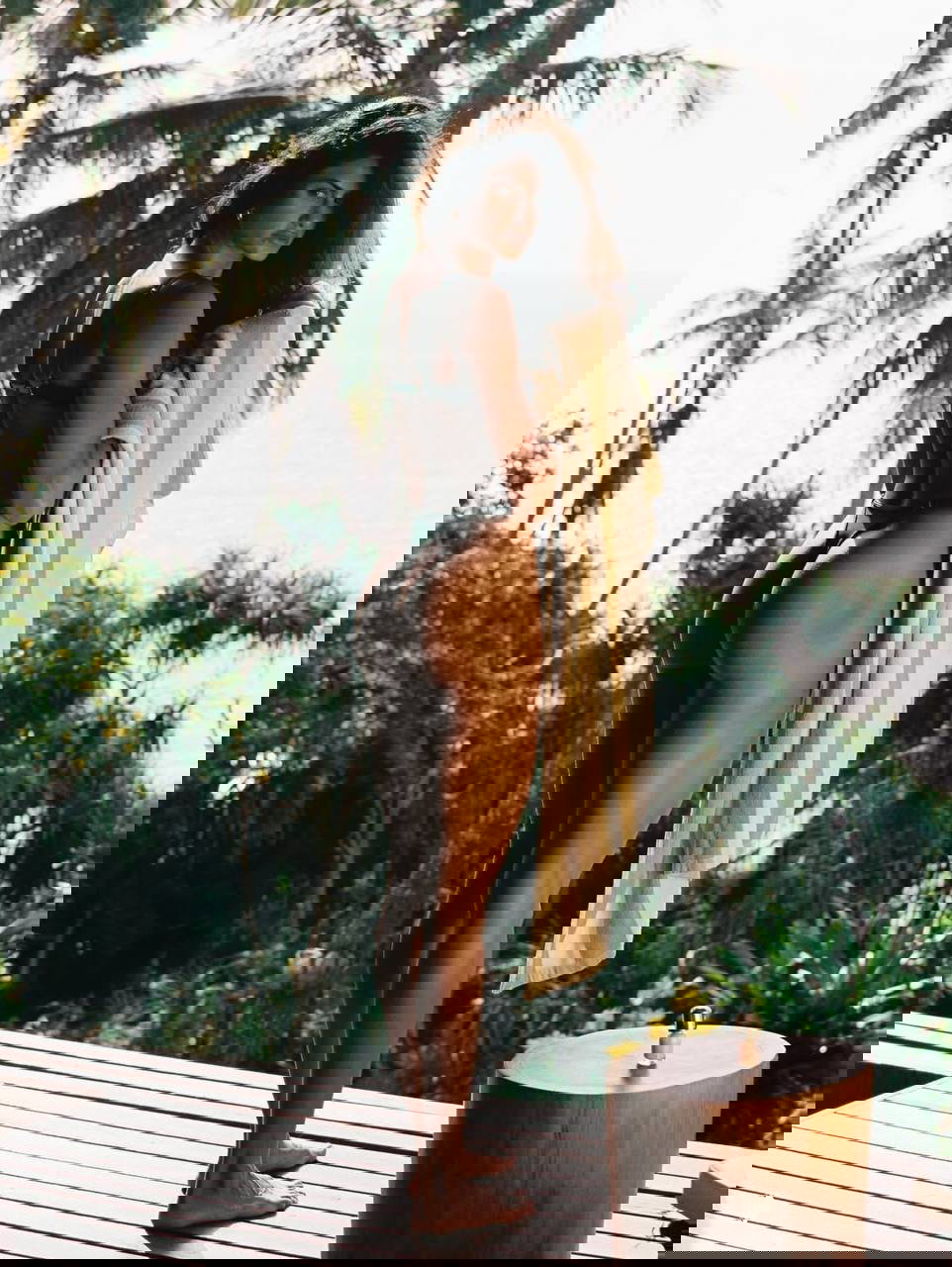 Модель стоит на террасе возле тропических растений с полотенцем, виднеется море на заднем плане, солнечный контражурный свет.