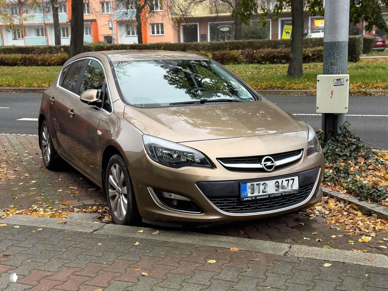 Бежевый Opel Astra, припаркованный у тротуара среди осенней листвы; вид в профиль и спереди показывает форму фар и бампера модели.