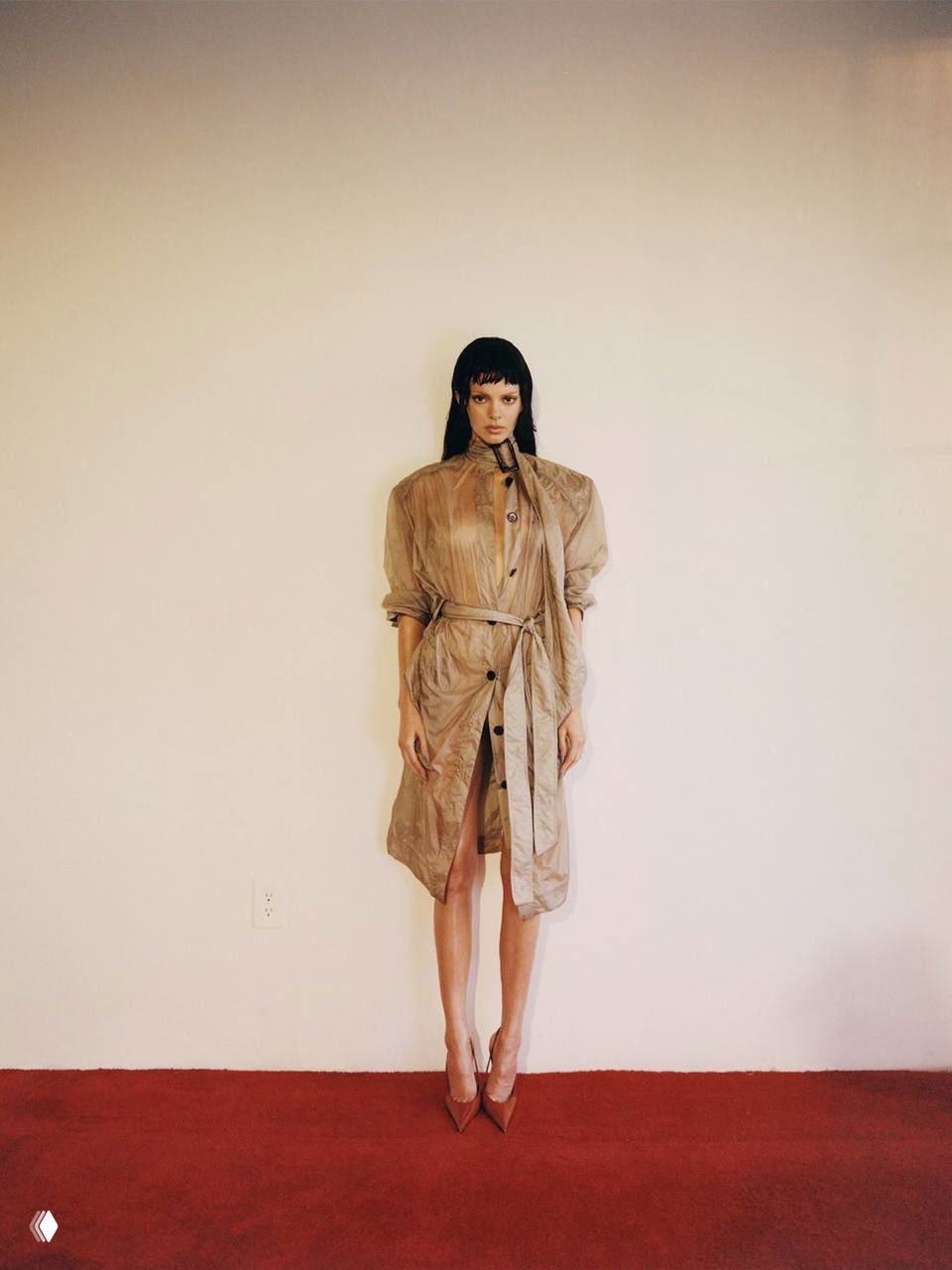Модель в лёгком плаще в полный рост стоит на красном покрытии в интерьере; акцент на силуэте, осмысленная простота образа и туфли на каблуке.
