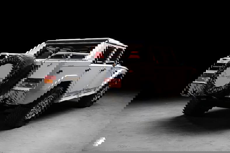 Задний ракурс белого Lamborghini LM002 с запасным колесом и открытой грузовой зоной, видны бампер, фонари и элементы рамы на чёрном фоне.