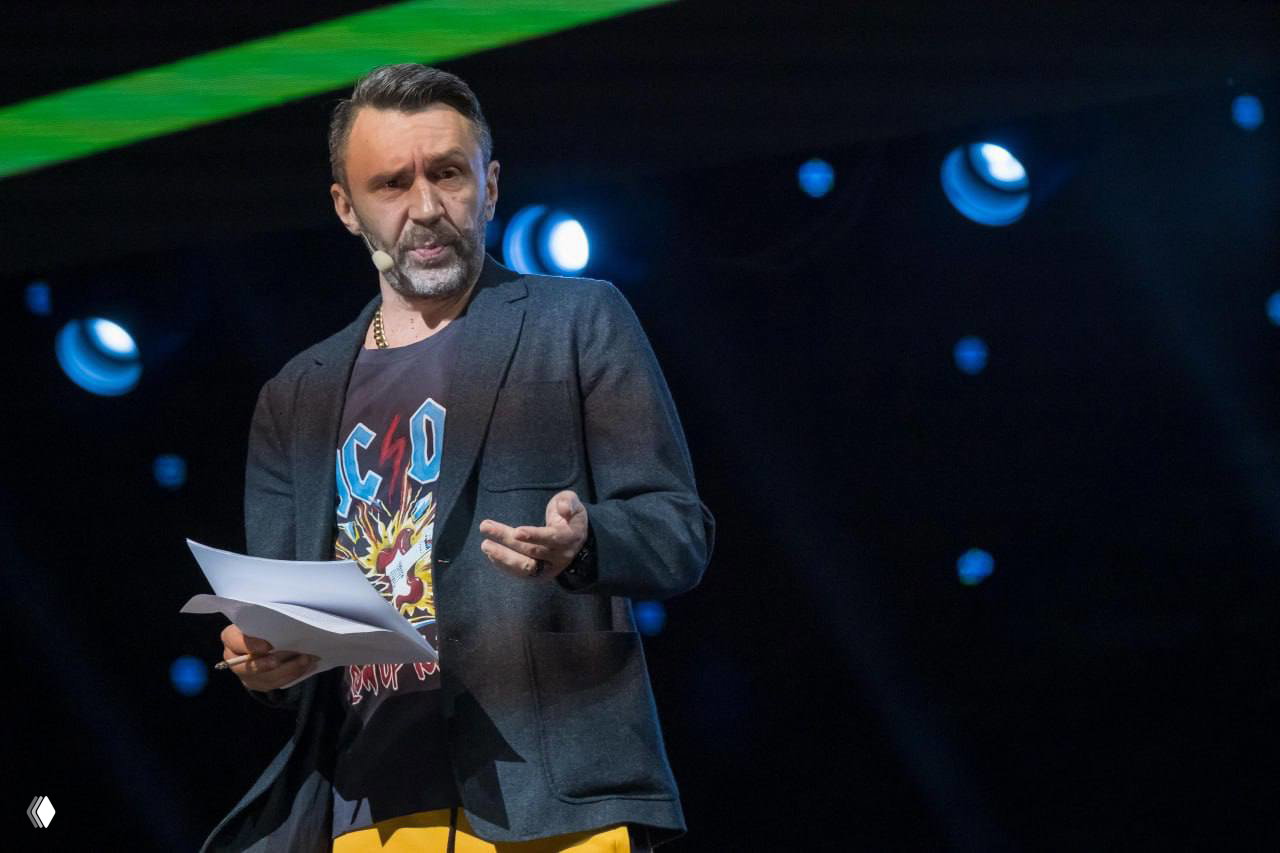 Мужчина на сцене в концертной обстановке держит листы и выступает в микрофон; драматичная подсветка и сценическое оформление, кадр выглядит динамично.