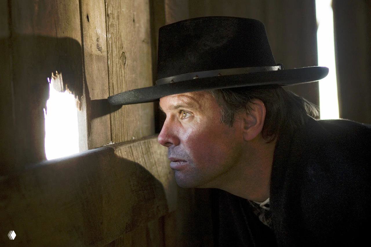 Крупный профильный портрет персонажа в шляпе, выглядывающего из деревянного строения: свет и тени подчёркивают напряжённое выражение лица.