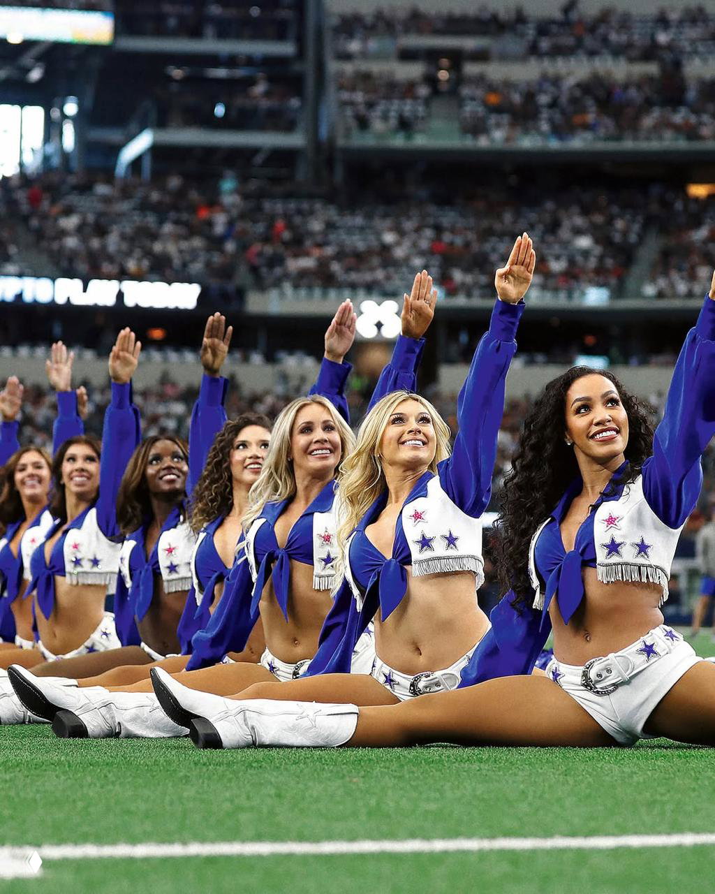 Группа чирлидерш Dallas Cowboys выстроена в ряд на поле стадиона в одинаковых синих костюмах и белых сапогах перед трибунами.