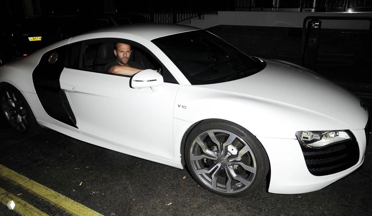 Мужчина сидит за рулём белого спорткара Audi R8 ночью на городской улице; видна эмблема V10 на крыле и переднее колесо.