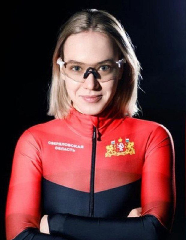 Конькобежка/шорт-трекистка в красном костюме с гербом региона и защитными очками, студийный портрет спортсменки на темном фоне.