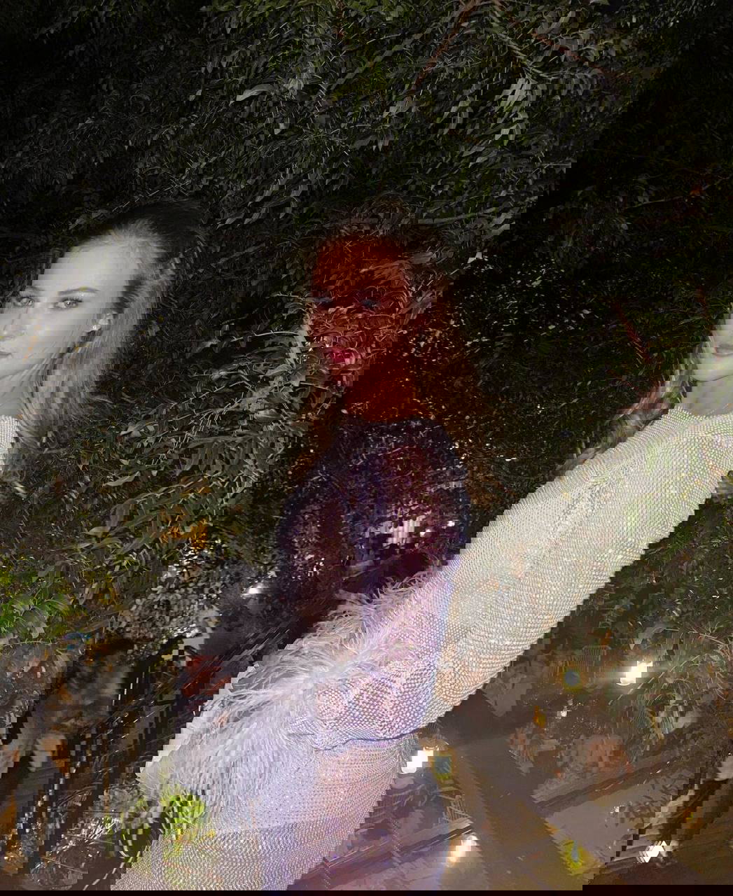 Женщина в вечернем платье с перьевыми рукавами на террасе между зеленью; светская обстановка с огнями и гостями внизу, портретная фотография с атмосферой мероприятия.