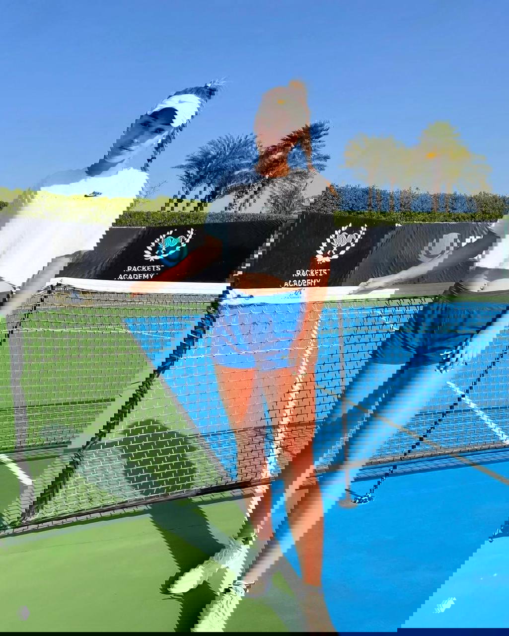 Молодая спортсменка позирует у сетки на корте в белой футболке и синих шортах; открытый тренировочный корт с пальмами на заднем плане и дневным солнечным светом.