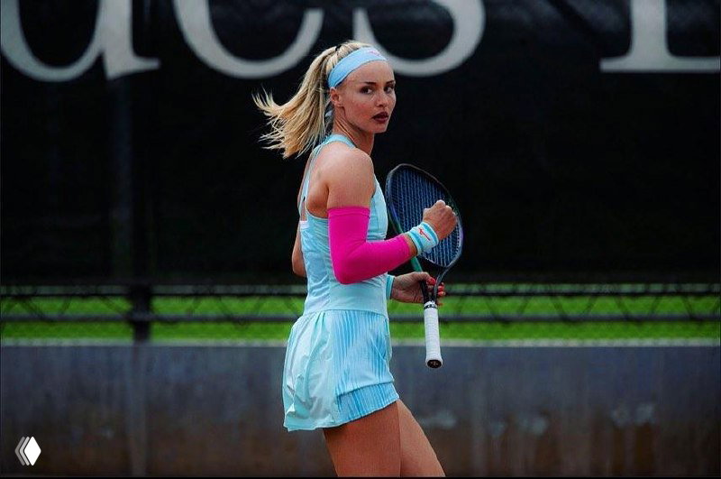 Теннисистка в голубом комплекте с повязкой и ярким розовым компрессионным рукавом, сосредоточенный взгляд во время матча; кадр с корта, эмоции и движение.