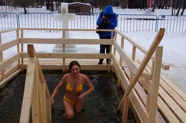 Девушка в жёлтом купальнике поднимается по ступеням деревянной проруби на фоне снега и ограждения; уличное крещенское купание.