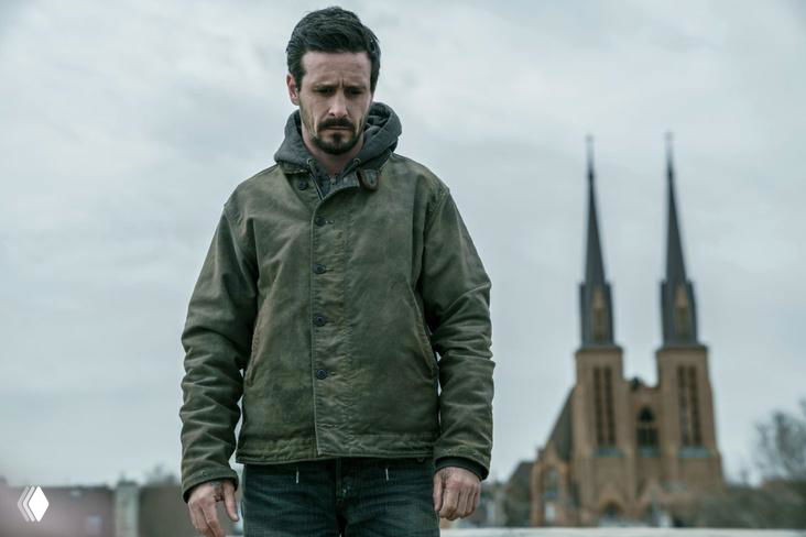 Кадр с Джеймсом Рэнсоном в тёмной куртке на фоне городской сцены с виднеющимися церковными шпилями, сцена из кинопроекта