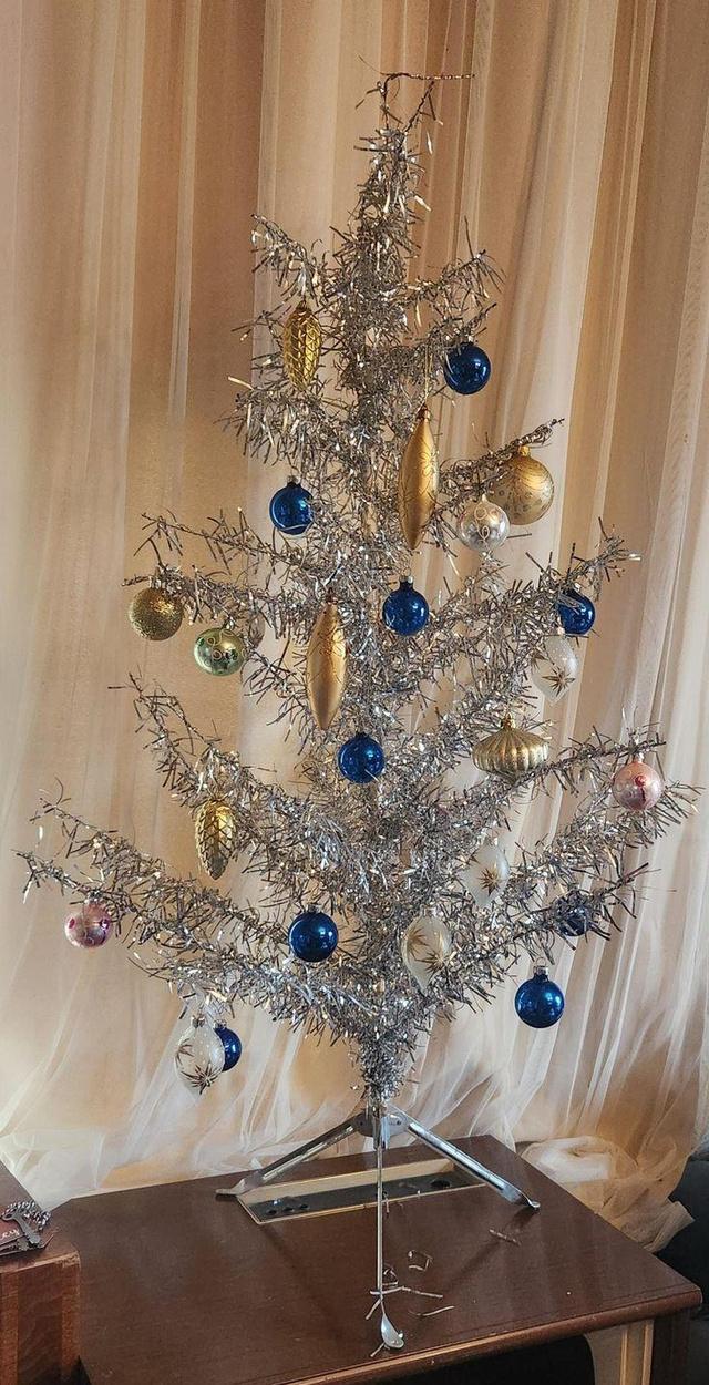 Металлическая серебристая ёлка на столе, украшенная синими и золотыми шарами; интерьерная фотография с простым фоном, типично для эпохи.