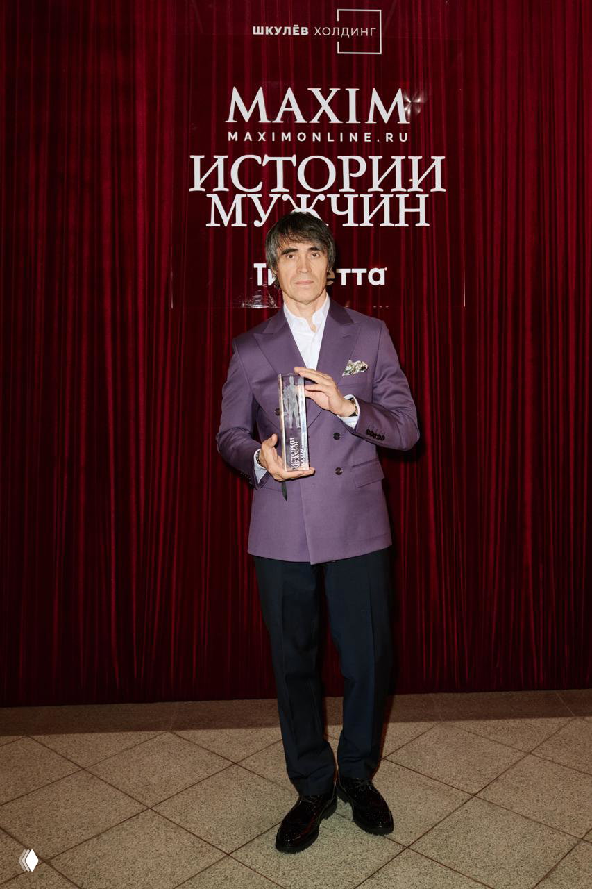 Мужчина в фиолетовом пиджаке позирует с хрустальной статуэткой на фоне брендовой декорации мероприятия «Истории мужчин».