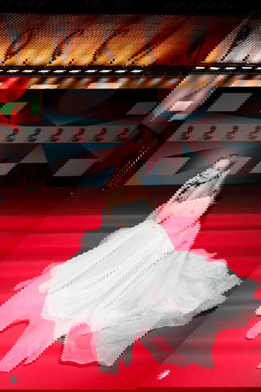 Селебрити в белоснежном пышном платье позирует на ступенях красной дорожки фестиваля на фоне баннеров и декораций мероприятия.