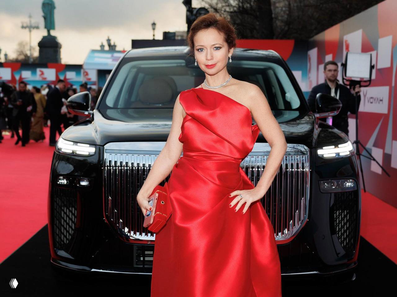 Актриса в атласном красном платье позирует на красной дорожке перед чёрным премиальным автомобилем-спонсором и фотозоной фестиваля.