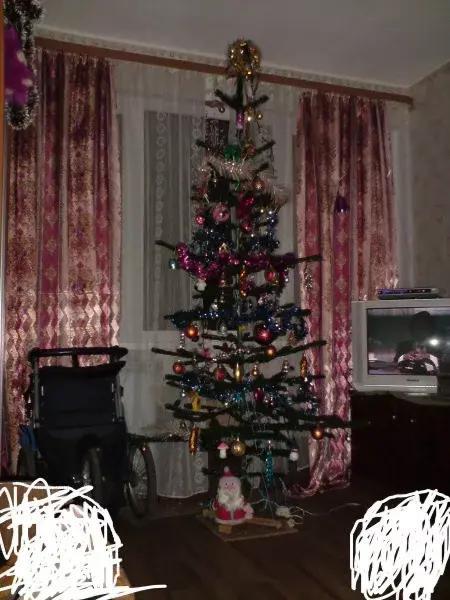 Живая ёлка в углу комнаты с гирляндами и игрушками; рядом видна инвалидная коляска и старый телевизор — реальный интерьер 1990-х годов.