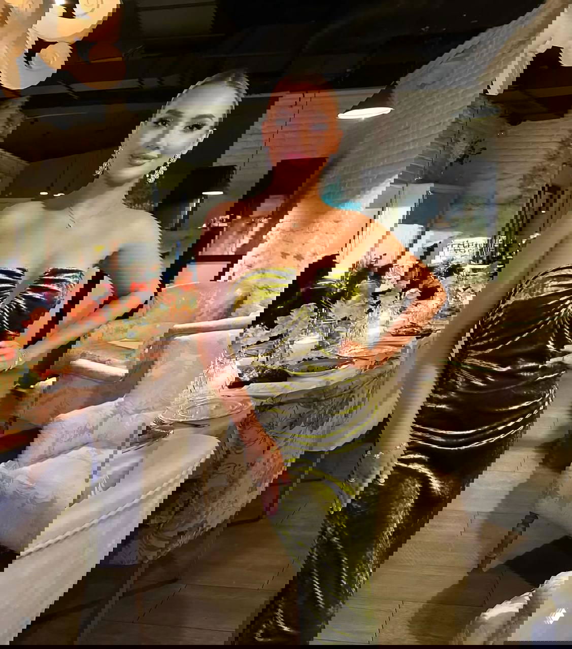 Женщина в оливково‑бархатном платье позирует у сервированного праздничного стола в интерьере ресторана, видны свечи и декор.