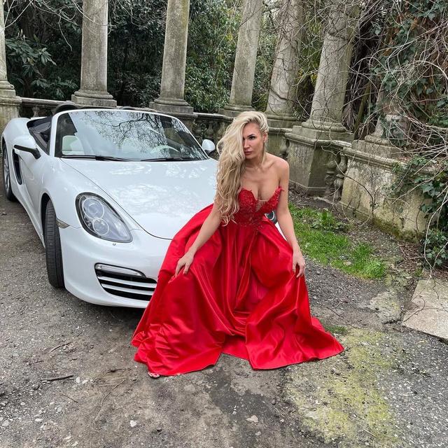 Женщина в длинном красном вечернем платье сидит рядом с белым кабриолетом у каменной ограды и колонн, поза для гламурной фотосессии на открытом воздухе.