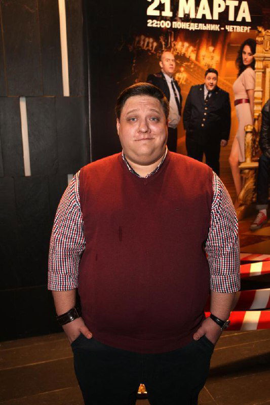 Роман Попов в повседневной одежде на мероприятии — полный рост актера на фоне афиши сериала «Полицейский с Рублевки», настроение красной дорожки.